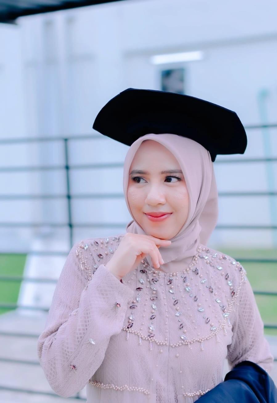 Ariantika smiling at the camera, wearing a cream hijab, round eyeglasses, and a white Genomic Science Day T-shirt with a light-blue lanyard, raising her right hand in a peace sign at an indoor research event.